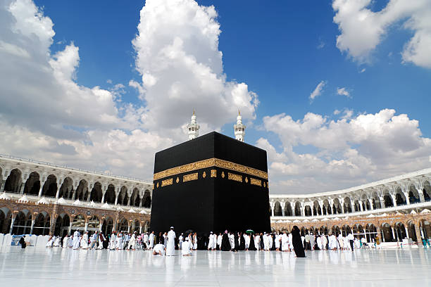Gambar menunjukkan Ka'bah, sebuah struktur berbentuk kubus besar berwarna hitam yang terletak di tengah Masjidil Haram di Mekah, Arab Saudi. Ka'bah dihiasi dengan kain bersulam emas yang dikenal sebagai Kiswah. Di sekitar Ka'bah, terdapat banyak orang yang mengenakan pakaian putih, sedang melakukan Tawaf, yaitu berjalan mengelilingi Ka'bah searah jarum jam. Arsitektur masjid menampilkan banyak lengkungan dan menara. Langit di atasnya sebagian berawan dengan beberapa bagian biru yang terlihat.