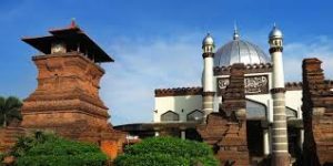 Kompleks Masjid Kudus berarsitektur perpaduan Jawa dan Islam, terdiri dari menara bata merah kuno dan bangunan masjid berkubah besar putih, dikelilingi tanaman hijau dengan latar langit biru cerah.